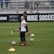 A person in sports attire stands on a soccer field, surrounded by several soccer balls and a carrier with bottles. The field is bordered by advertisements for Nike Soccer and app stores. Several people sit in bleachers in the background.
