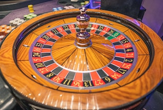 A roulette wheel with alternating red and black numbered pockets on a wooden platform. The wheel contains a small white ball resting in one of the pockets, indicating gameplay. Surrounding the wheel are stacks of colorful poker chips, suggesting a casino environment.
