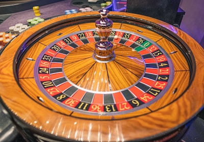 A roulette wheel with alternating red and black numbered pockets on a wooden platform. The wheel contains a small white ball resting in one of the pockets, indicating gameplay. Surrounding the wheel are stacks of colorful poker chips, suggesting a casino environment.