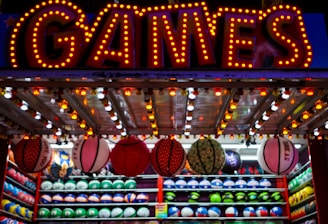A brightly lit carnival game stall with a large 'GAMES' sign at the top surrounded by red and yellow lights. Various sports balls, including basketballs and soccer balls, are hanging from the ceiling and neatly arranged on shelves below.