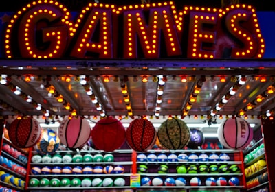 A brightly lit carnival game stall with a large 'GAMES' sign at the top surrounded by red and yellow lights. Various sports balls, including basketballs and soccer balls, are hanging from the ceiling and neatly arranged on shelves below.